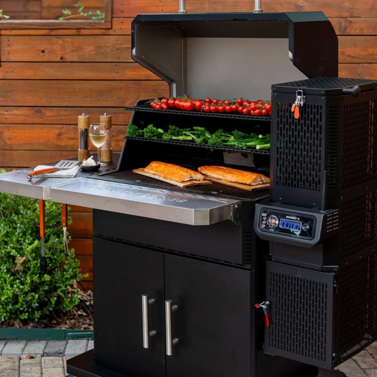 Masterbuilt Gravity Series XT digital charcoal smoker in a backyard setting, featuring an open lid with multiple cooking racks holding salmon, vegetables, mushrooms, and bread, alongside a side firebox unit with digital temperature controls and stainless steel work surface.